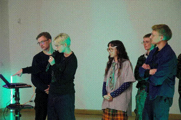 A group of five people are standing and looking at a computer. One of them is talking into the microphone and pointing at the screen as they talk about what happened during the workshop.